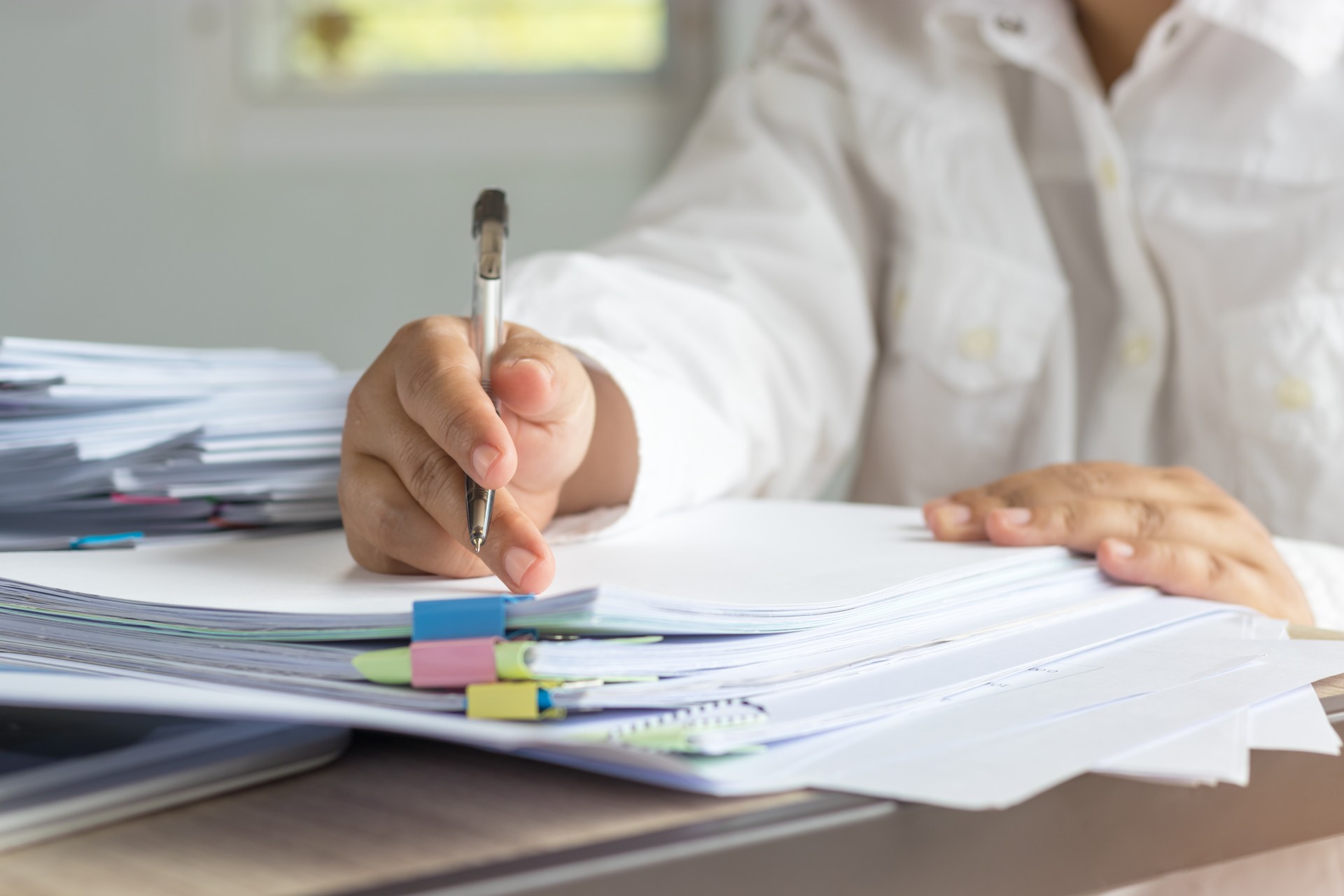 Teacher hand is holding pen for checking student homework assignments on desk in school. Unfinished paperwork stacked in archive with color paper and binder paper clips. Education and business concept